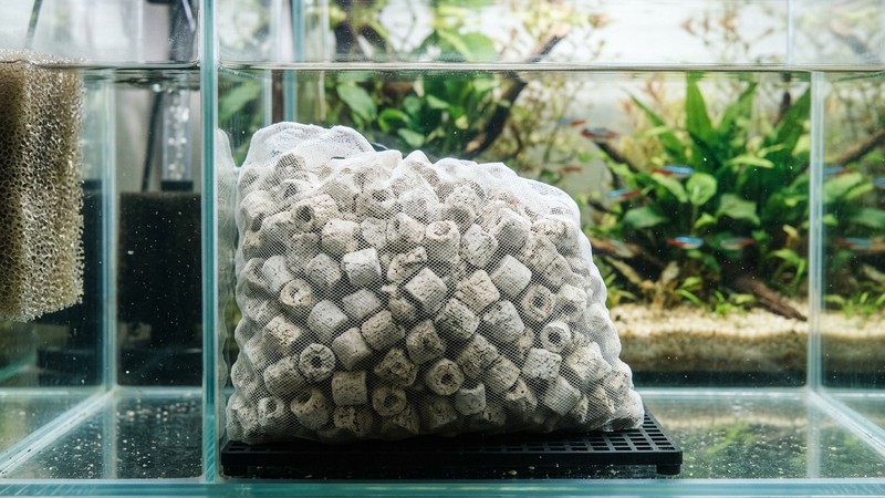 una bolsa de malla llena de canutillos cerámicos en reposo dentro del compartimento de un filtro de acuario, textura porosa y limpia, junto al cristal del tanque