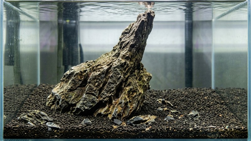 una roca de aquascaping de base ancha y punta estrecha, con textura rugosa y aristas irregulares, apoyada sobre sustrato oscuro dentro de un acuario de agua clara